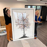 Two people holding a large framed artwork of a tree with birds in an indoor setting.